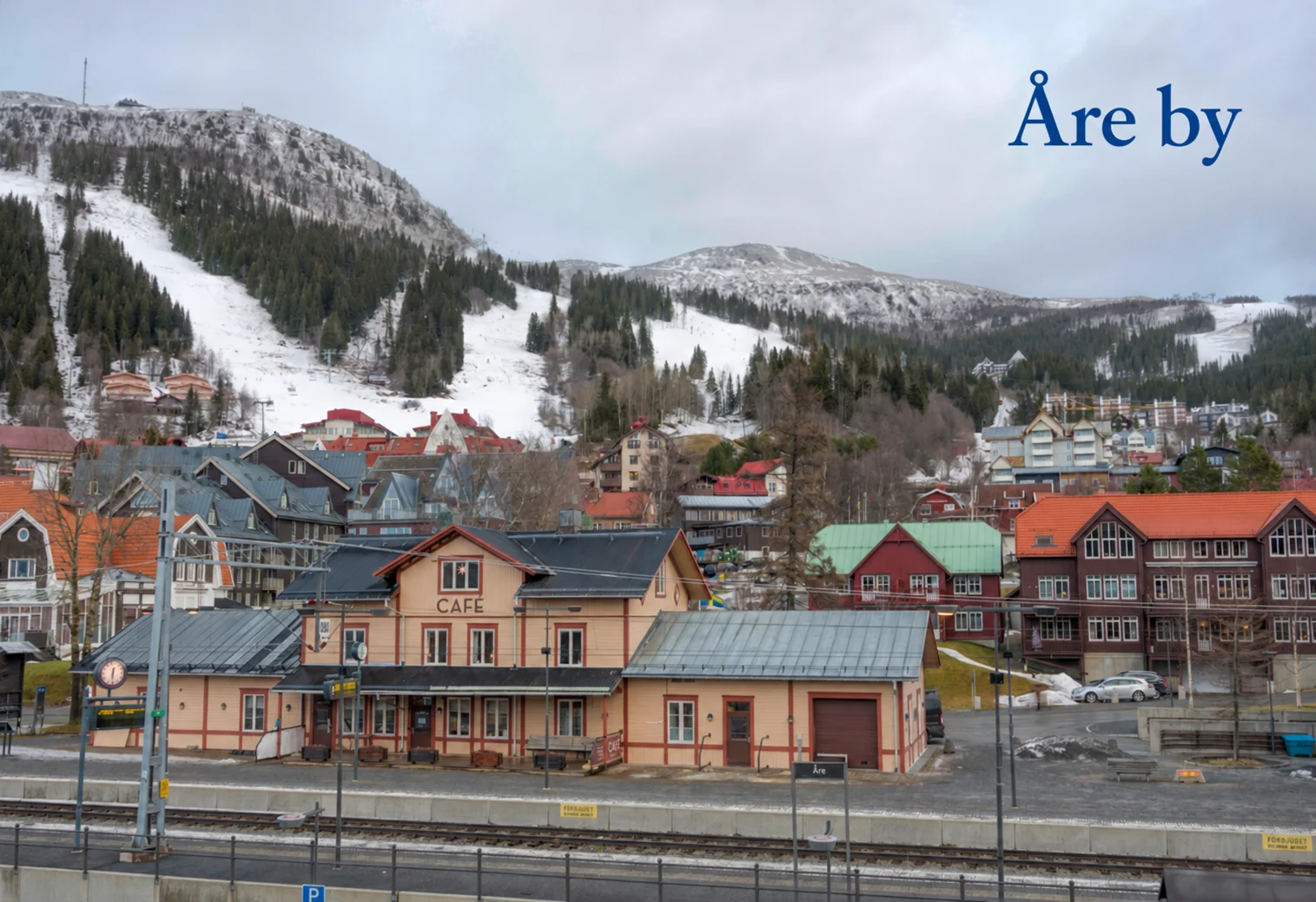 Åre by med järnvägsstationen, hus och snötäckta skidbackar. Texten Åre by är inlagd i himlen.