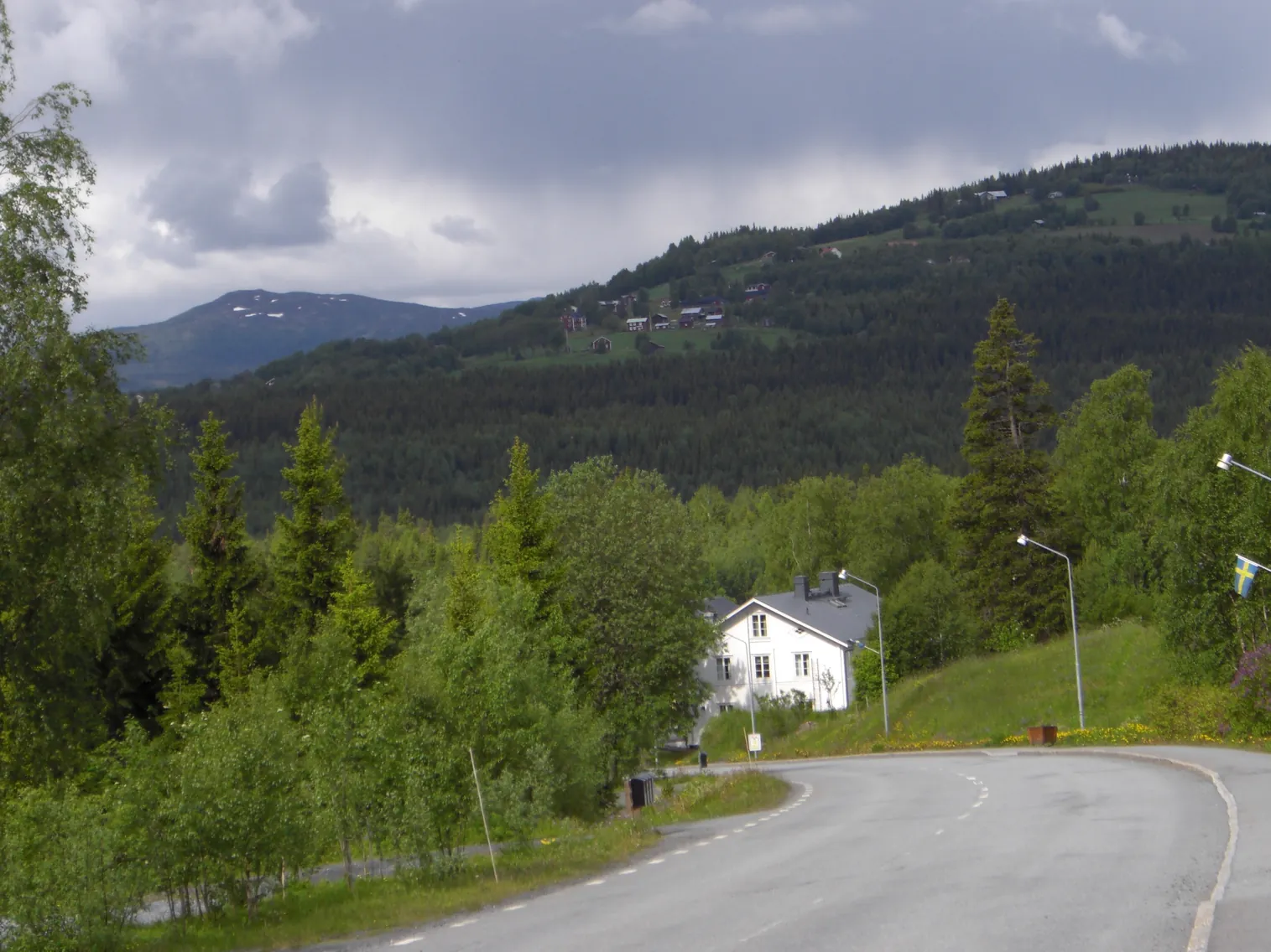 Väg genom Undersåker med ett vitt hus, björkskog och fjäll i bakgrunden.